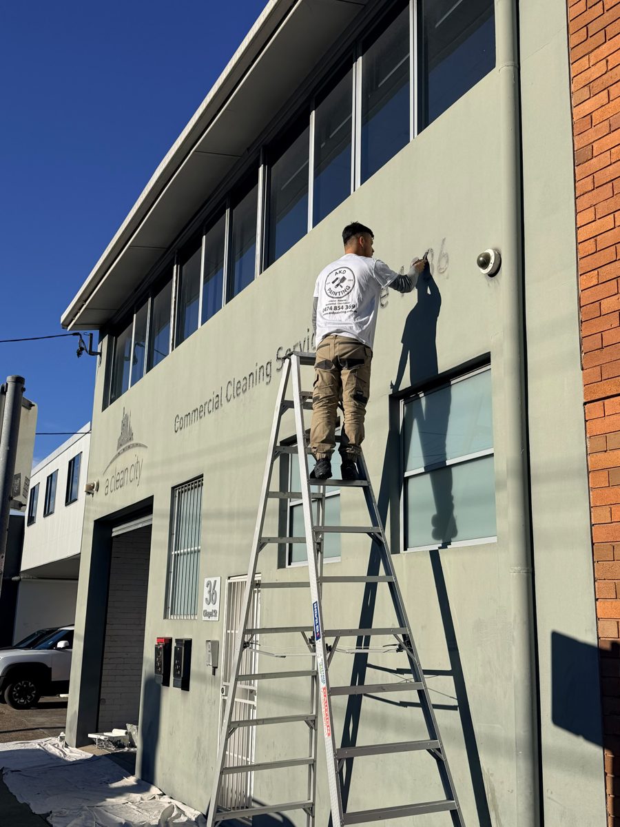 AKD painter on a platform ladder removing old painted signage from a two-storey commercial building in Sydney