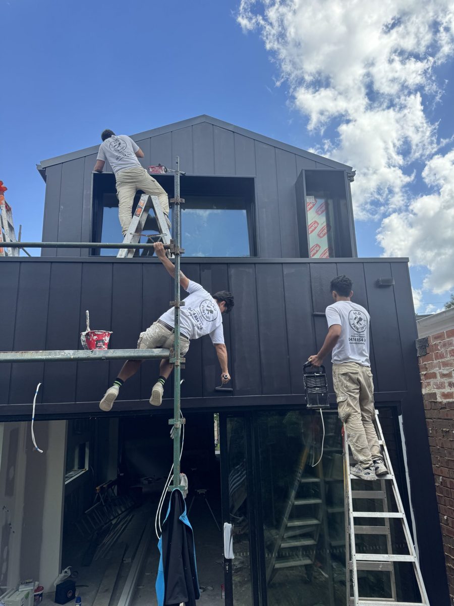 Three AKD painters on ladders painting the black vertical cladding of a two-storey rear extension in Leichhardt