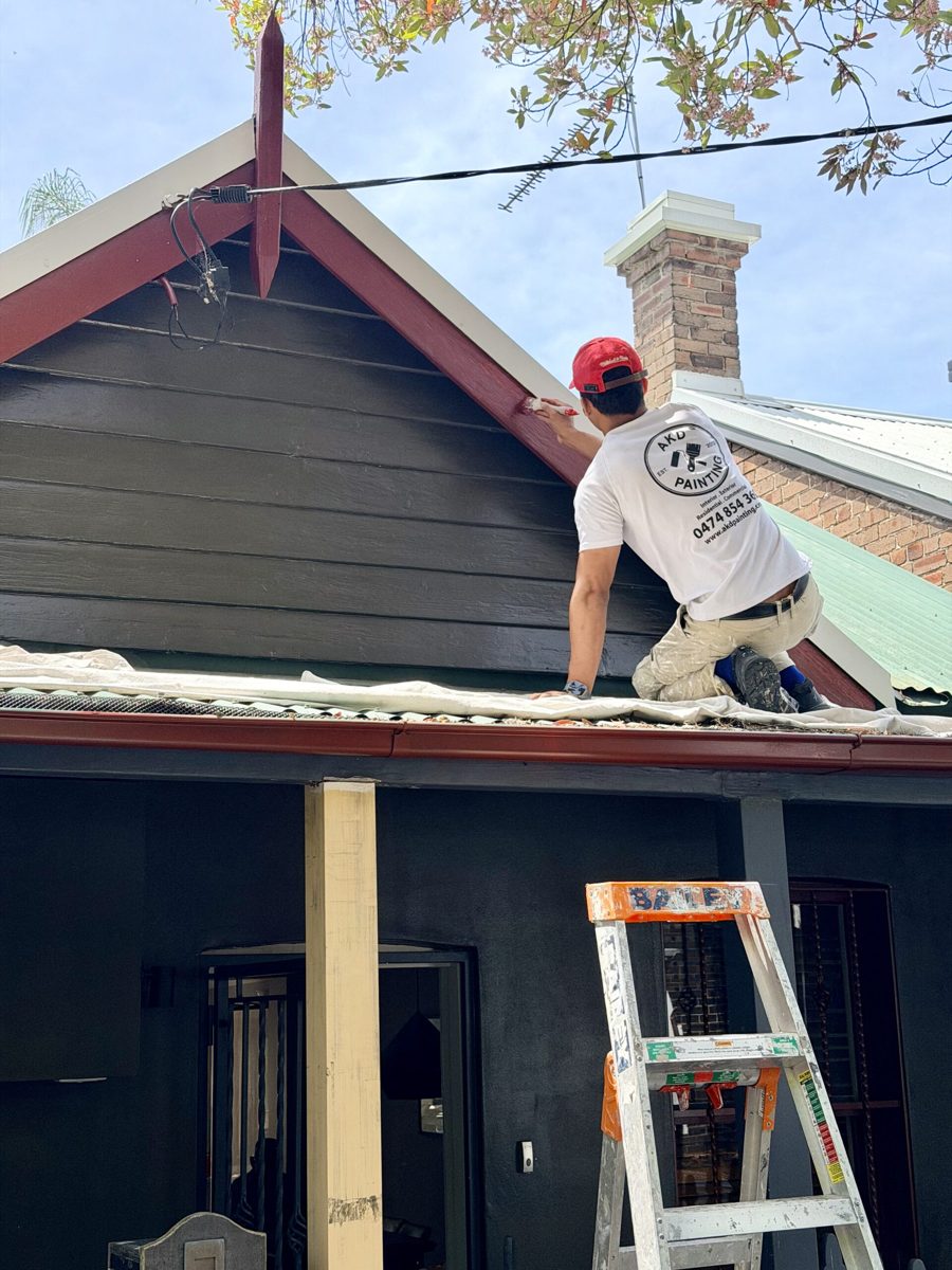 AKD painter on the verandah roof painting the red barge board of a weatherboard cottage gable in Newtown