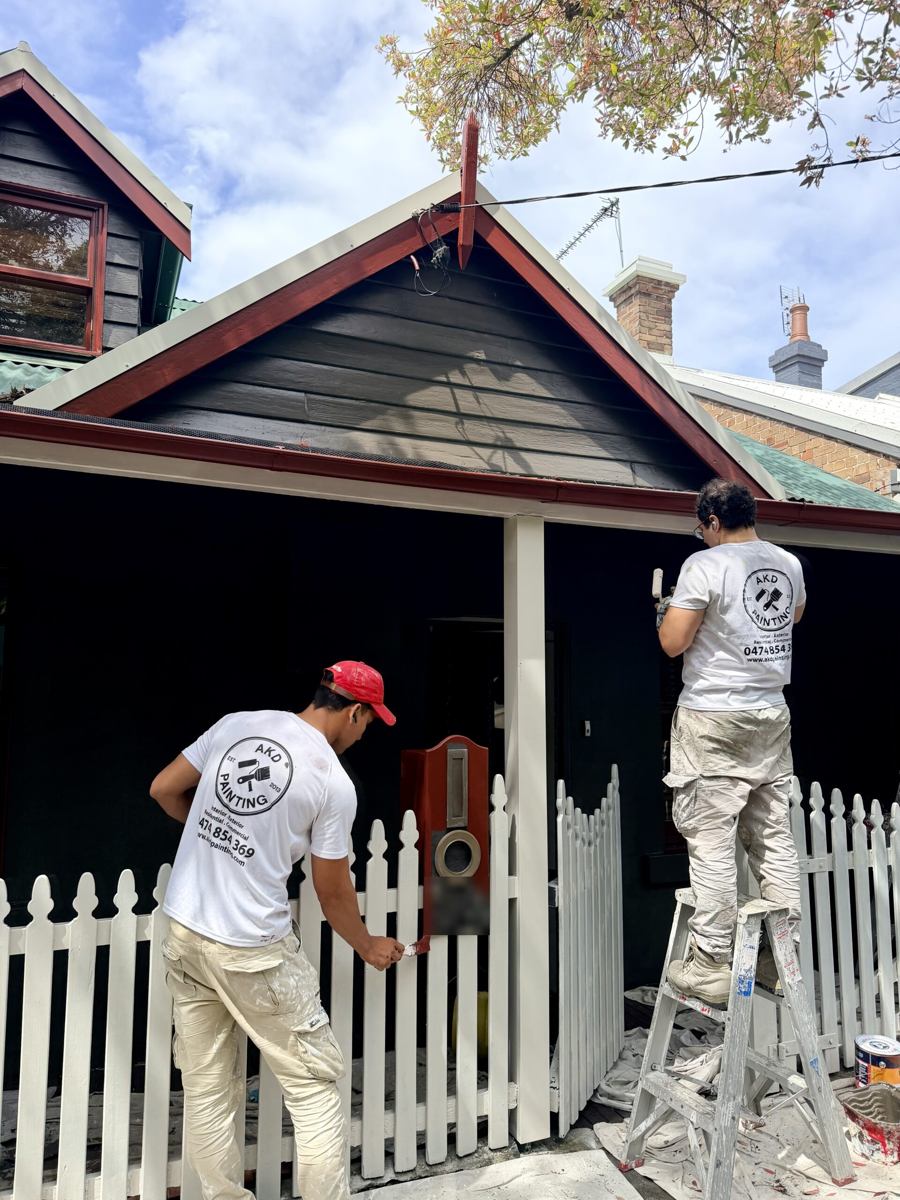 Two AKD painters repainting the picket fence and verandah post of a charcoal weatherboard cottage in Newtown