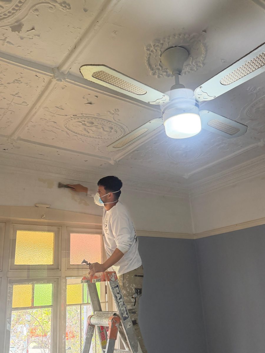 AKD painter on a step ladder scraping the edge of a pressed metal ceiling in a Five Dock bedroom
