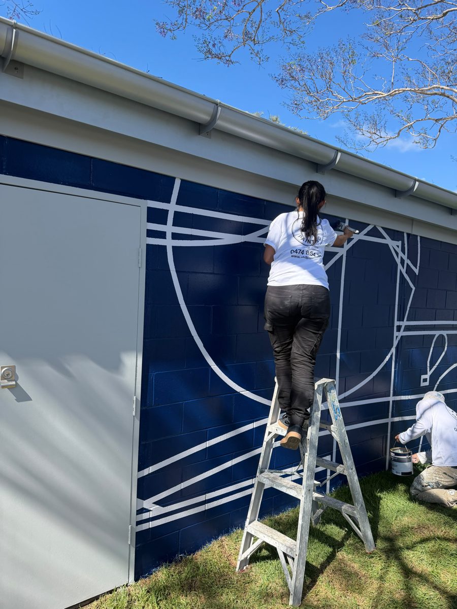 AKD painter on a step ladder touching up white lines by brush on a navy amenities block in Marrickville