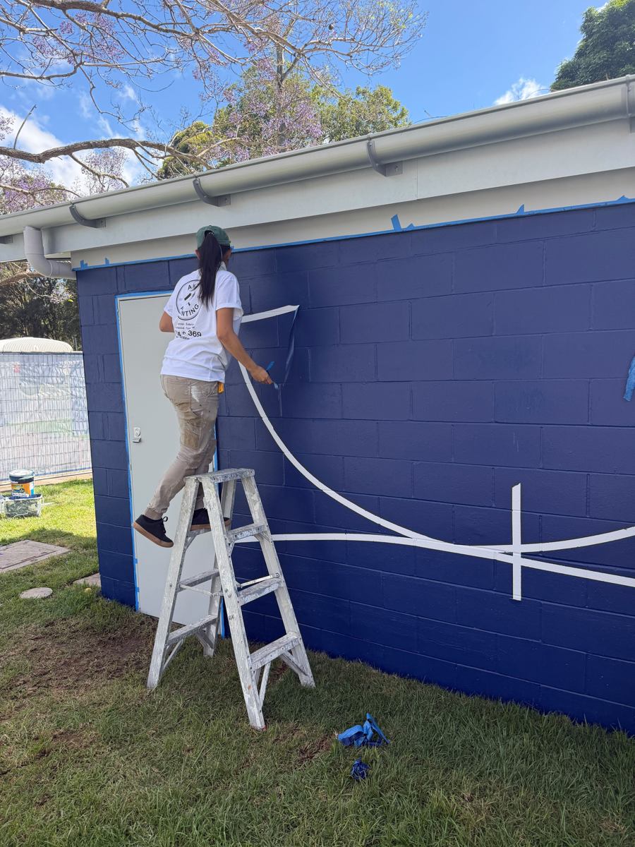 AKD painter pulling masking tape from a navy wall to reveal white curved lines on a Marrickville amenities block