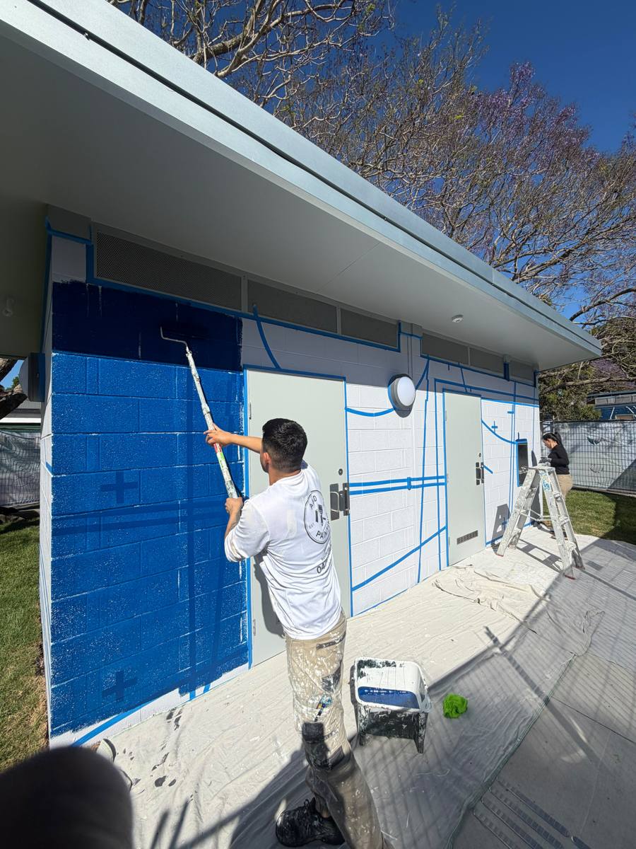 AKD painter rolling navy paint over taped linework on the front wall of a Marrickville amenities block