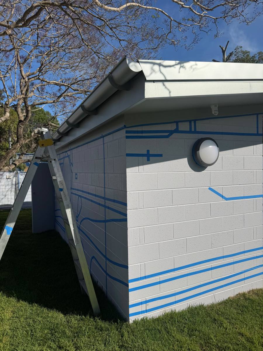 End wall of a Marrickville park amenities block in pale grey with the linework design masked out in blue tape